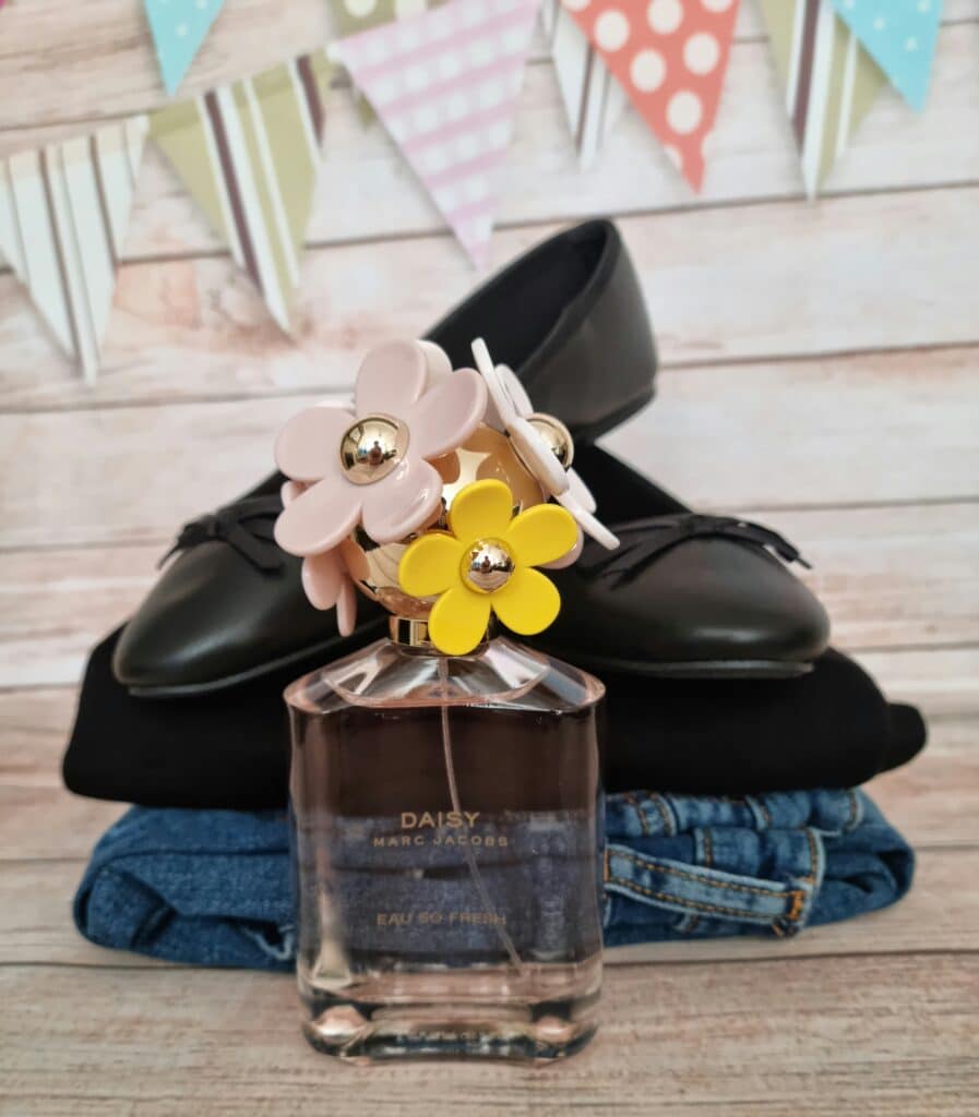 A bottle of Daisy by Marc Jacobs perfume sits in front of folded black and denim clothing, with a pair of black shoes and colourful bunting in the background—perfect for celebrating Mother’s Day with Very.