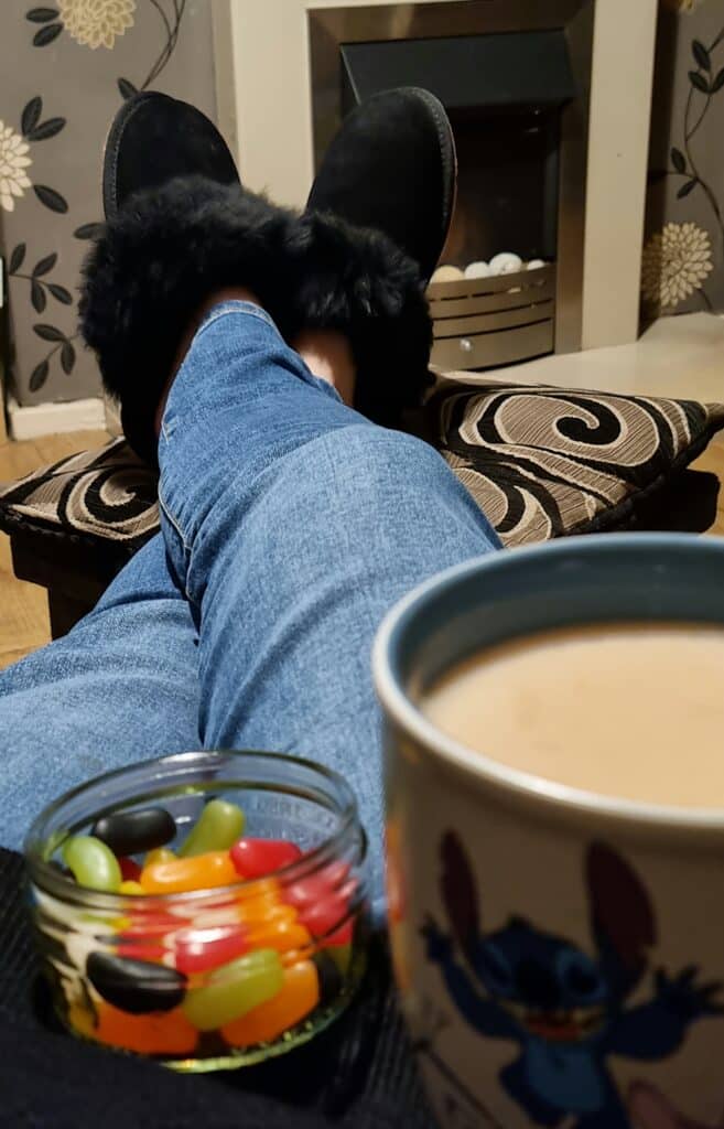 A person wearing blue jeans and black slippers lounges with feet up, enjoying a Stitch mug and a bowl of colourful jelly beans by the fireplace—perfect for relaxing on Mother’s Day with Very.