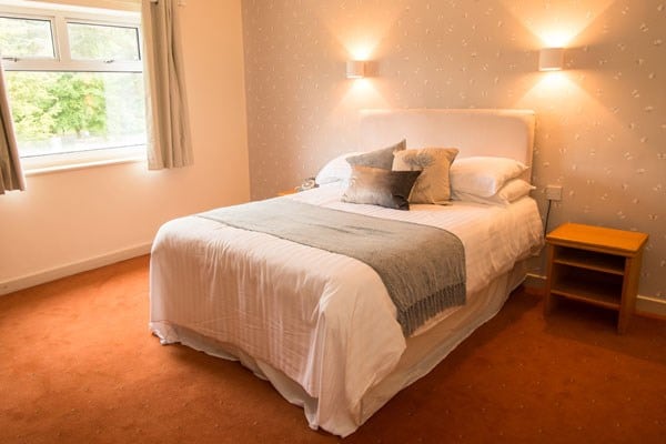 A cosy bedroom with a double bed, white bedding, grey throw, and decorative cushions—perfect for Valentine's Day. Two wall lights above the bed, a wooden bedside table, and a window with curtains letting in natural light.
