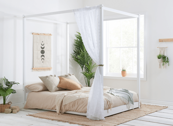 Bright bedroom features one of the 10 bed frames guaranteed to impress guests—a white canopy bed draped in sheer curtains, neutral bedding, hanging plants, a woven rug, wall art, and a large window streaming natural light.