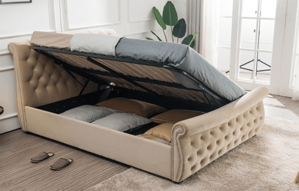 One of 10 bed frames guaranteed to impress guests, this beige upholstered storage bed lifts up to reveal spacious storage for pillows and blankets. Slippers rest nearby, whilst a green plant sits by the window in the background.