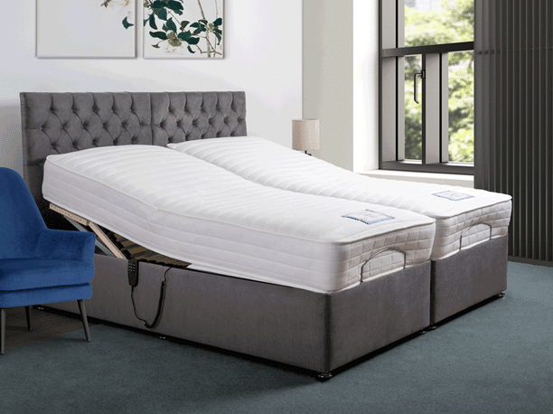 A modern bedroom with a grey upholstered adjustable bed—one of 10 bed frames guaranteed to impress guests—features split sections with raised headrests, a blue chair, a lamp, wall art, and a large window framing views of trees outside.