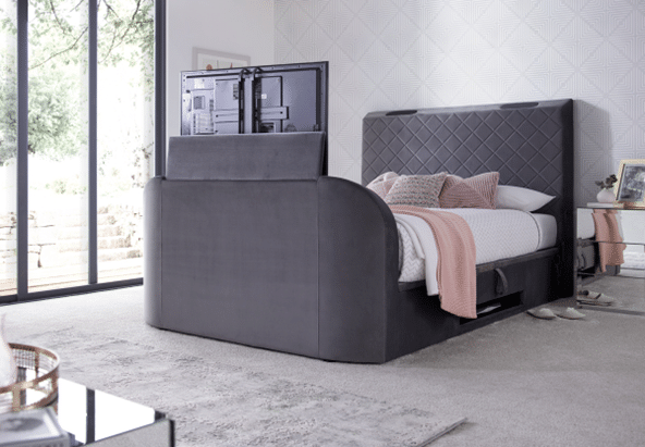A modern bedroom with a large grey upholstered bed featuring a built-in TV lift at the footboard—one of 10 bed frames guaranteed to impress guests. Light carpet, soft pink bedding accents, and large windows complete the inviting space.