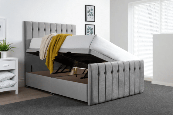 A grey upholstered bed with lift-up mattress reveals under-bed storage—one of 10 bed frames guaranteed to impress guests. The room features white walls, framed art, a bedside table with shelves, and a yellow blanket draped over the bed.