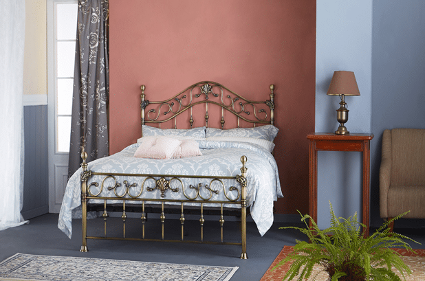 A bedroom with a decorative metal bed frame—one of 10 Bed frames Guaranteed to Impress Guests—white and light blue bedding, a wooden bedside table with a lamp, beige armchair, patterned curtains, a plant, and soft pink and blue walls.