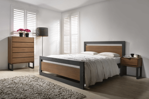 Modern bedroom with a wooden bed frame—one of 10 bed frames guaranteed to impress guests—plus matching bedside table and chest of drawers. White bedding, black floor lamp, potted flowers, and a textured rug complete the space bathed in sunlight from plantation shutters.