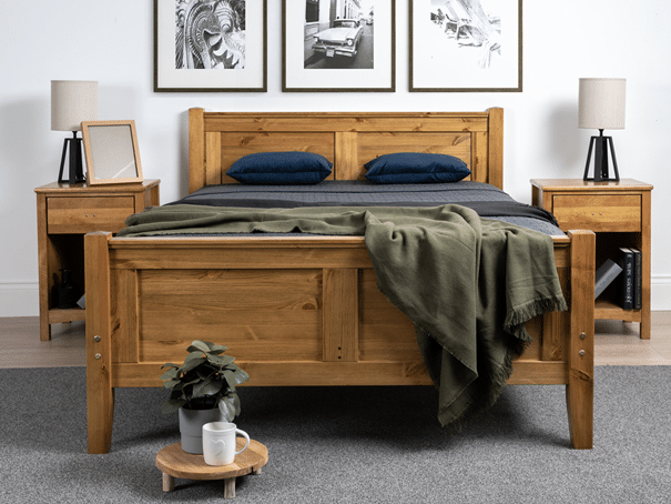 A wooden bed from our 10 Bed Frames Guaranteed to Impress Guests features dark bedding, matching bedside tables with lamps, and framed art above. A plant on a round stand adds charm at the foot of the bed atop a grey carpeted floor.