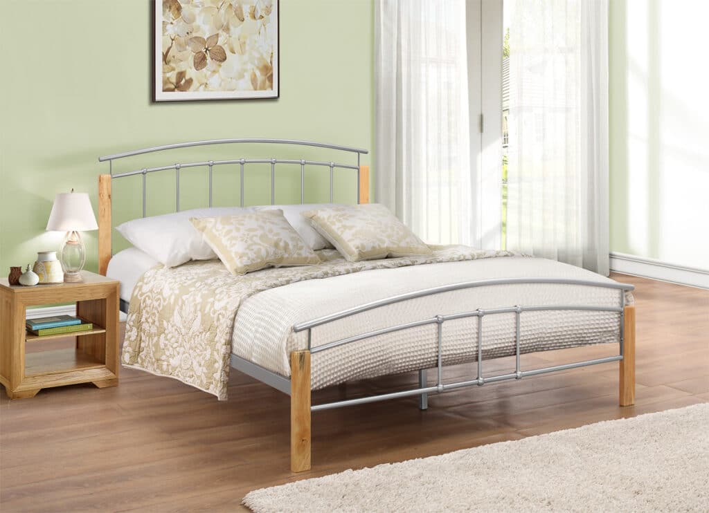 A neatly made bed with a metal and wooden frame, inspired by the style of 10 bed frames guaranteed to impress guests, stands in a bright bedroom with light green walls, a bedside table, lamp, and decor. Sunlight streams through sheer curtains over large glass doors.