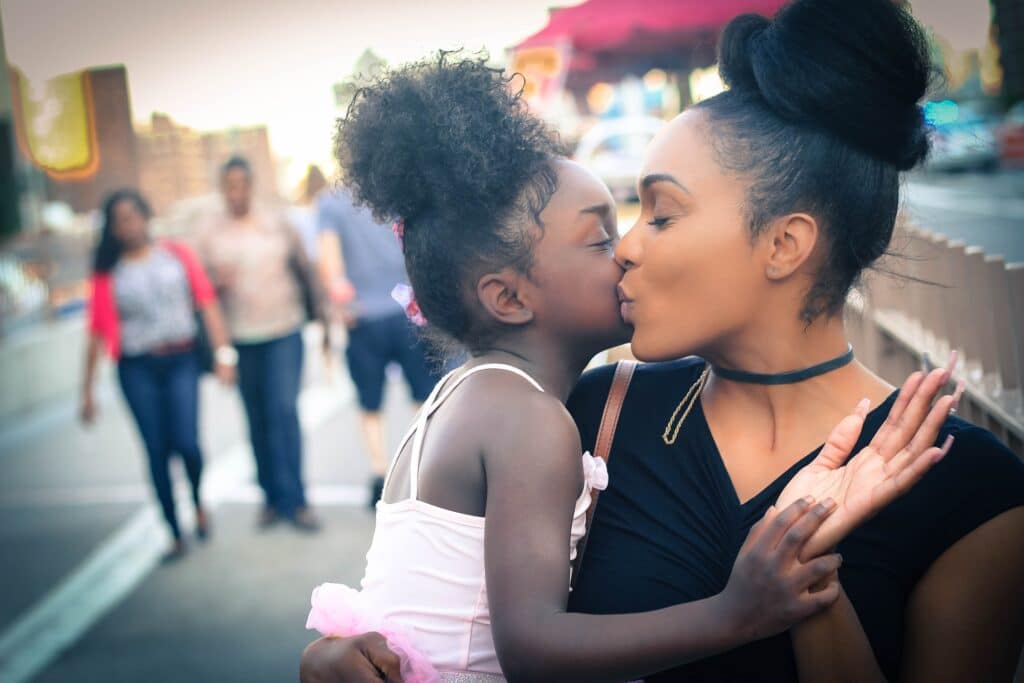 A young girl kisses her mother on the lips as they embrace on a city street, both smiling with eyes closed; people and buildings are visible in the blurred background.