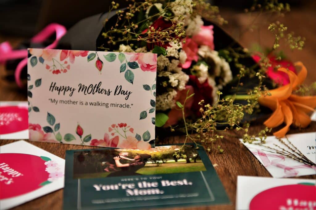 A bouquet of flowers and several Mother’s Day cards are spread on a wooden table. The main card reads, “Happy Mother’s Day. My mother is a walking miracle.” Celebrate Mother’s Day with very special cards and blooms.