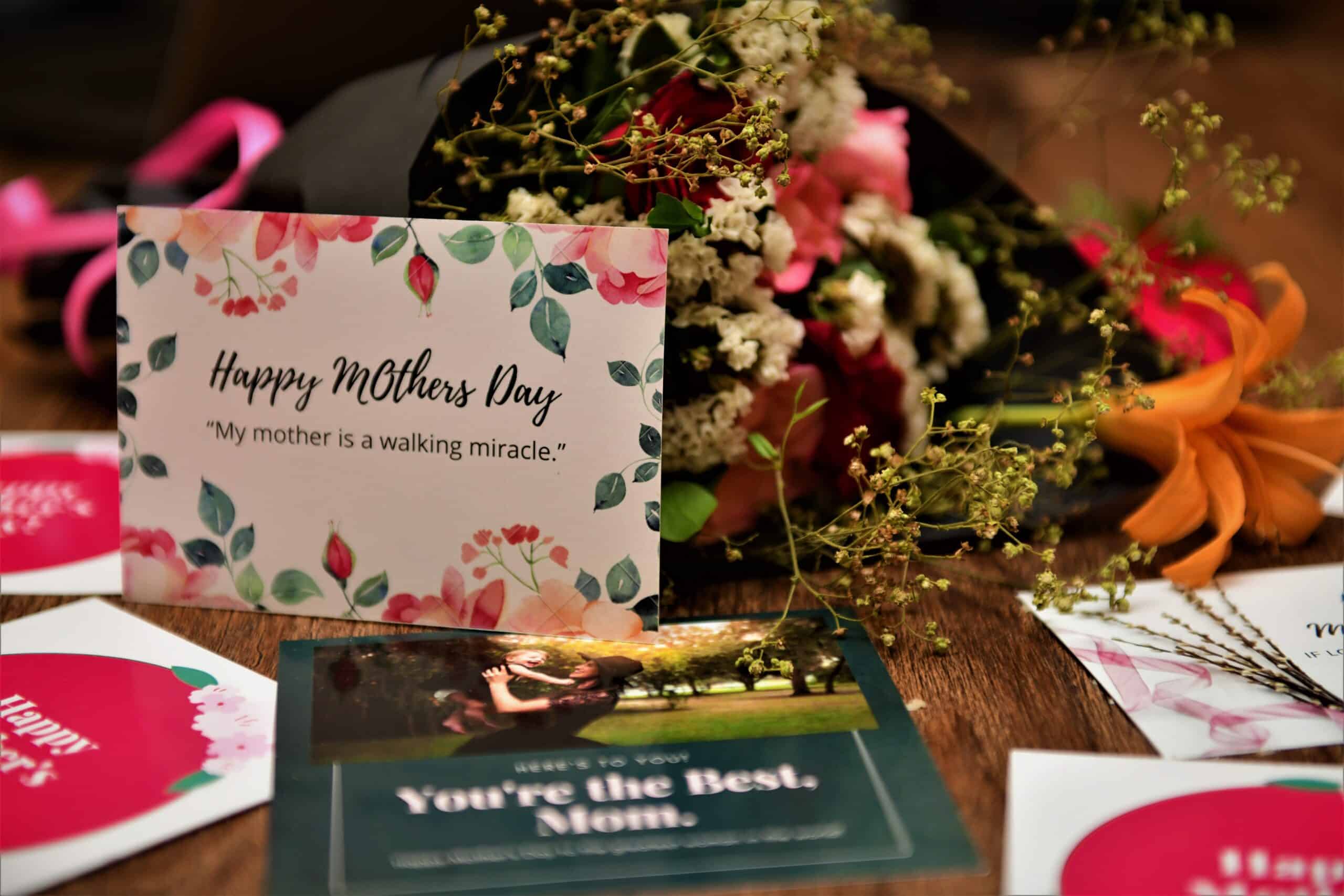 A bouquet of flowers and several Mother’s Day cards are spread on a wooden table. The main card reads, “Happy Mother’s Day. My mother is a walking miracle.” Celebrate Mother’s Day with very special cards and blooms.