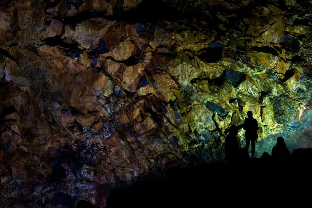 Three people stand in silhouette against a rugged, multicoloured cave wall illuminated by dramatic lighting—a true bucket list experience that highlights the textures and patterns of the rock surface.