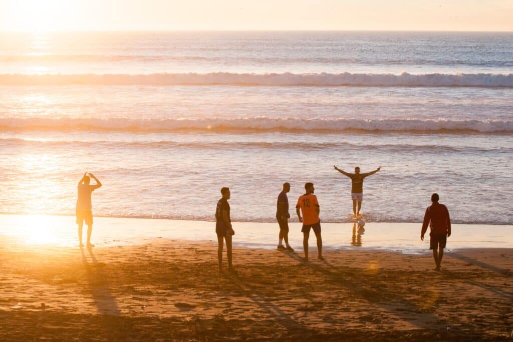 Six people stand and play on a sandy beach at sunset, with gentle waves in the background. Long shadows are cast by the setting sun, making this bucket list moment unforgettable as one person raises their arms near the water’s edge.