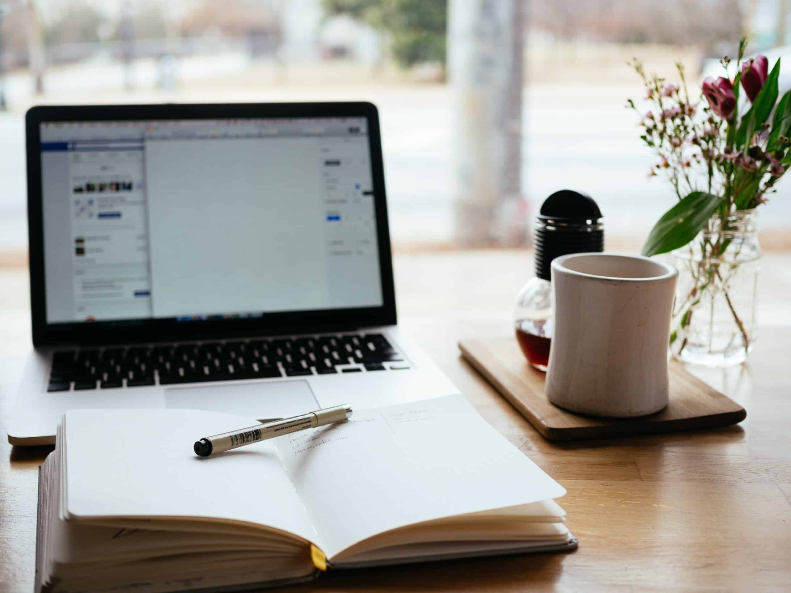 A laptop displaying a document sits on a wooden desk beside an open notebook with a pen, a white mug, a small pot, and a vase of flowers—an inviting setting for discovering tips for working from home, with sunlight streaming through the window.
