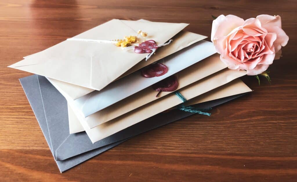 A stack of sealed envelopes, some with wax seals, lies on a wooden surface next to a pale pink rose—evoking nostalgia in contrast to modern e-cards.