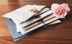 A stack of sealed envelopes, some with wax seals, lies on a wooden surface next to a pale pink rose—evoking nostalgia in contrast to modern e-cards.