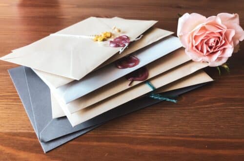 A stack of sealed envelopes, some with wax seals, lies on a wooden surface next to a pale pink rose—evoking nostalgia in contrast to modern e-cards.