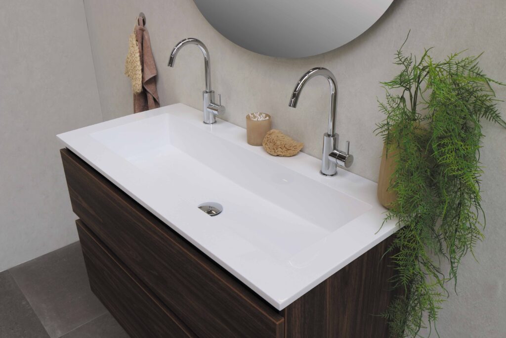 Modern bathroom basin with two chrome taps on a wooden vanity unit, perfect for small bathrooms. A round mirror, beige wall, hand towels, and a green potted plant highlight how 5 Types of Vanity Units That are a Perfect Fit for a Small Bathroom.