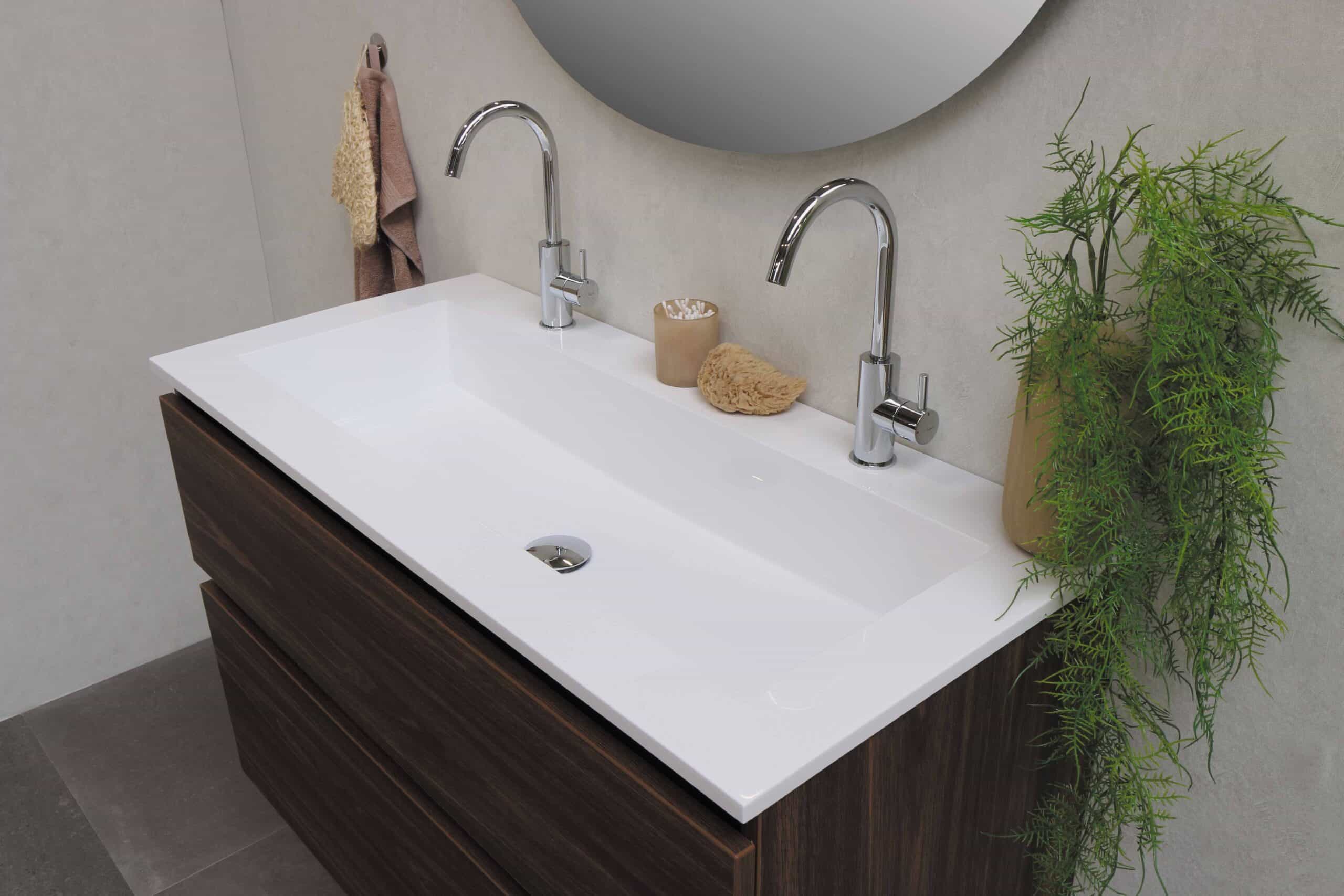Modern bathroom basin with two chrome taps on a wooden vanity unit, perfect for small bathrooms. A round mirror, beige wall, hand towels, and a green potted plant highlight how 5 Types of Vanity Units That are a Perfect Fit for a Small Bathroom.