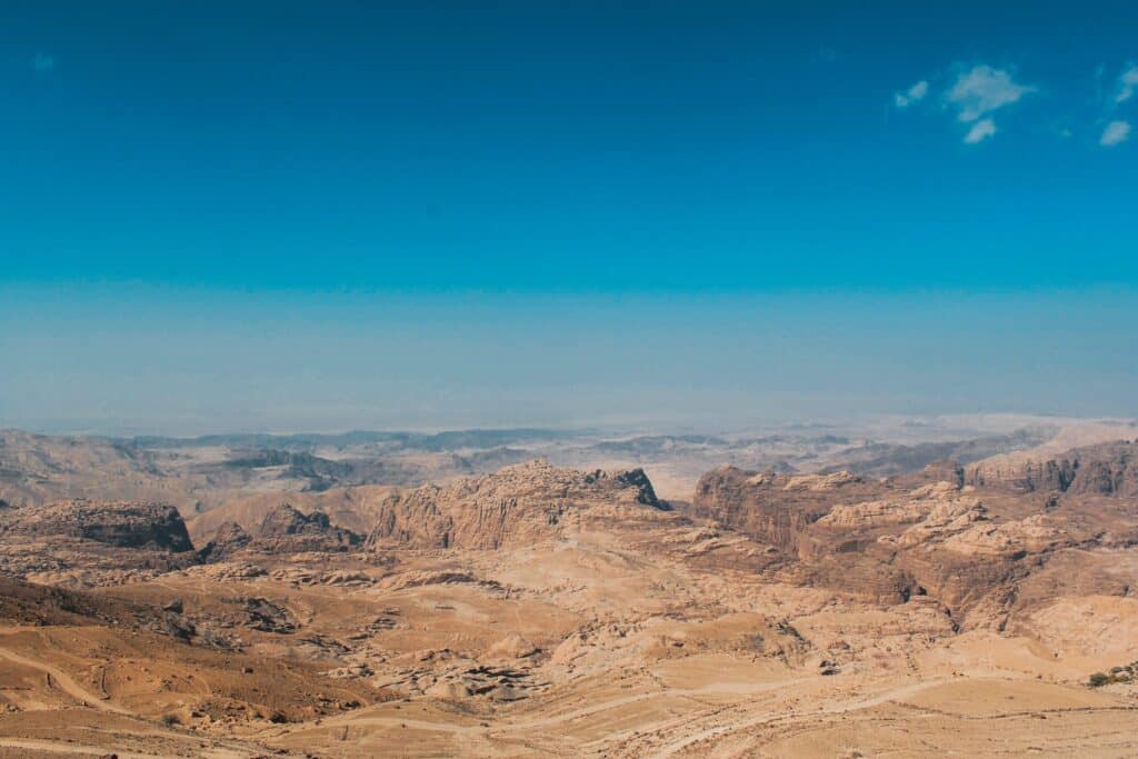 A vast, rocky desert landscape with rugged hills and valleys under a clear blue sky stretches into the distant horizon—a true bucket list destination for adventure seekers.