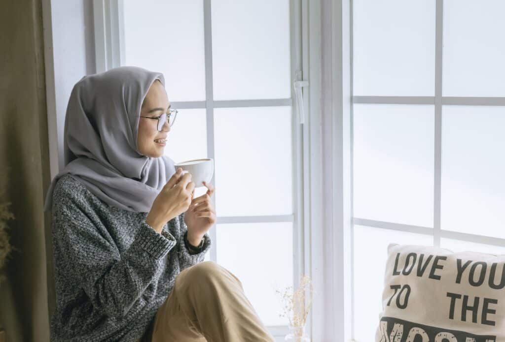 A mother wearing glasses and a grey hijab sits by a bright window, smiling and holding a white mug. A decorative cushion beside her reads "LOVE YOU TO THE MOON.