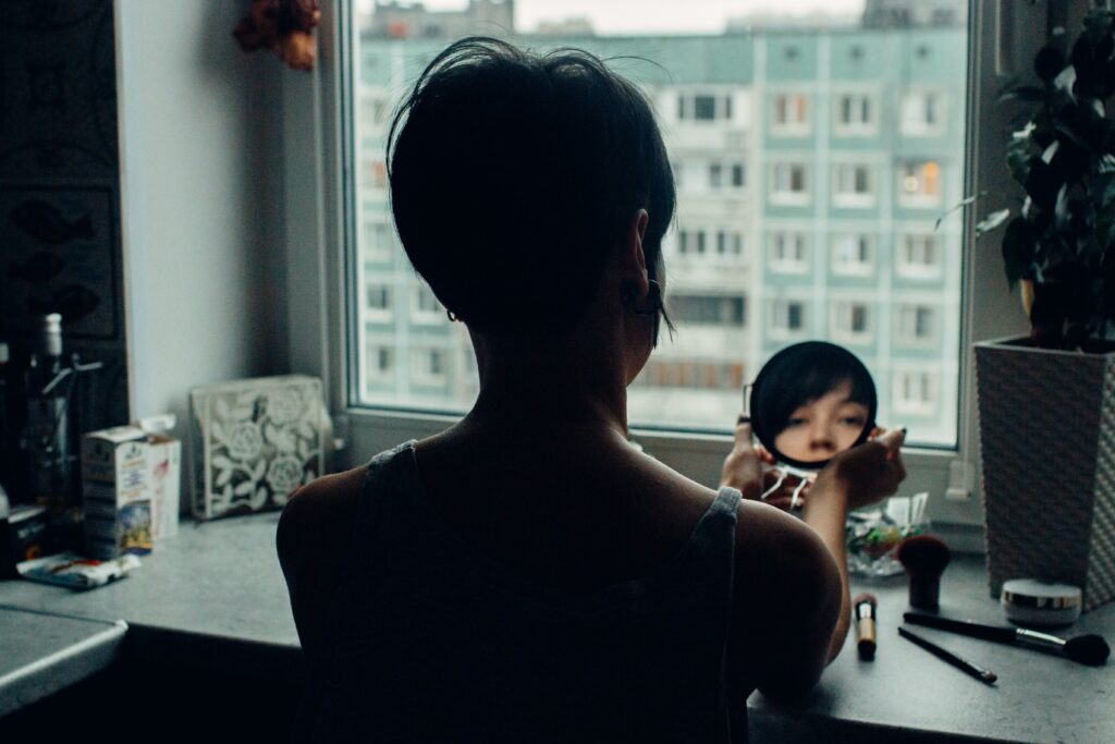 A person with short dark hair sits by a window, holding a small round mirror that reflects part of their face—perhaps checking for a common cause of skin cyst in women. Block of flats are visible outside, and make-up items are scattered on the table.
