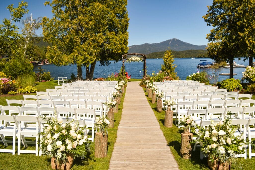 Rows of white chairs face a wooden wedding arch adorned with flowers, set on a lakeside lawn with mountain views—an idyllic scene that might make you wonder: are destination weddings really worth it? White floral arrangements line the aisle.