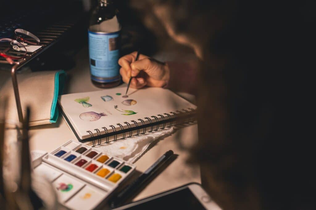 A person uses a small paintbrush to create colourful watercolour illustrations in a sketchbook, finding relaxation—one of four helpful ways to avoid parenting burnout—with a palette of paints, a closed pencil case, and a bottle on the desk nearby.