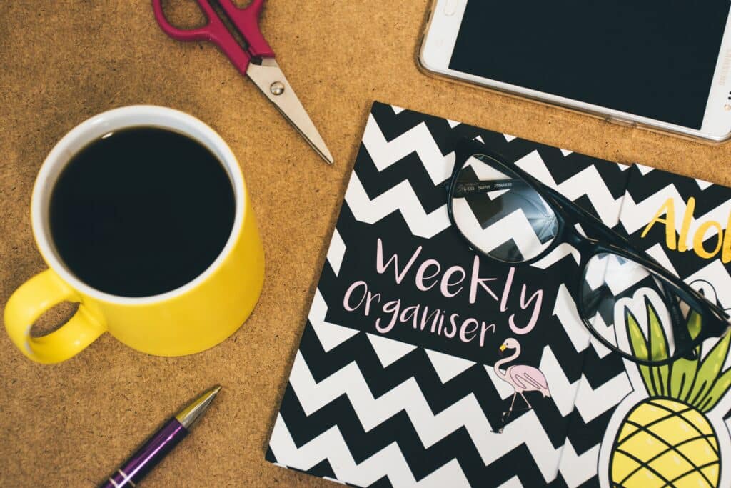 A yellow mug of black coffee, a pair of scissors, a tablet, pen, sunglasses, and a chevron-patterned weekly organiser notebook with a flamingo and pineapple design are on a brown surface—perfect for jotting down 4 Helpful Ways To Avoid Parenting Burnout.