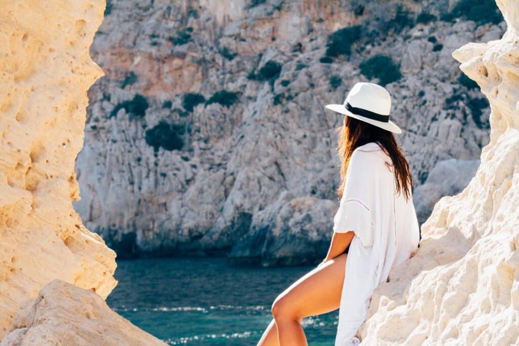A woman in a white hat and cover-up sits on rocky cliffs, looking out over blue water and sunlit stone formations—capturing that sense of awe you feel when going abroad for the first time: what you need to know for an unforgettable journey.