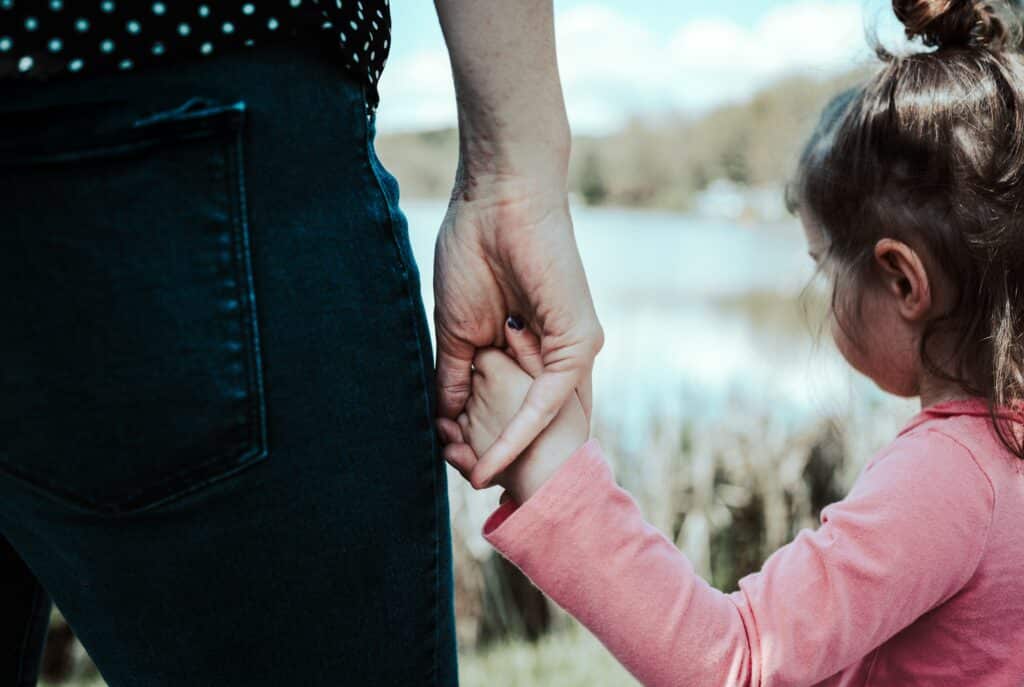 An adult and a young girl hold hands outdoors by a lake. The adult, in a perfect Mother’s Day outfit of dark jeans and a spotted black shirt, joins the girl in pink, highlighting their special connection and care.