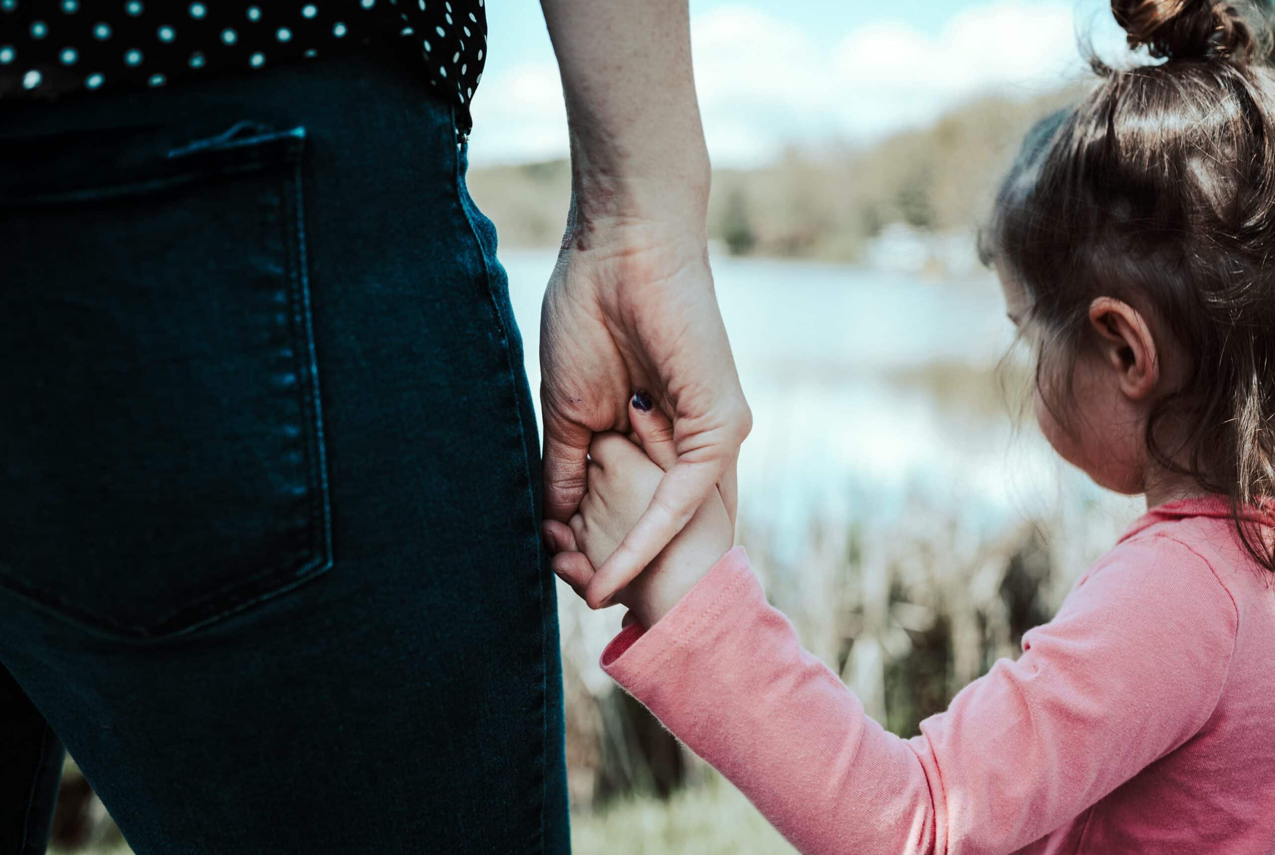 An adult and a young girl hold hands outdoors by a lake. The adult, in a perfect Mother’s Day outfit of dark jeans and a spotted black shirt, joins the girl in pink, highlighting their special connection and care.