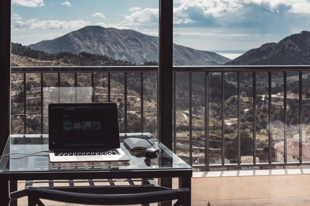 A laptop sits on a glass table next to sunglasses and a mouse, with a mountain and valley view visible through large windows—perfect for those seeking the world’s best locations for remote working.