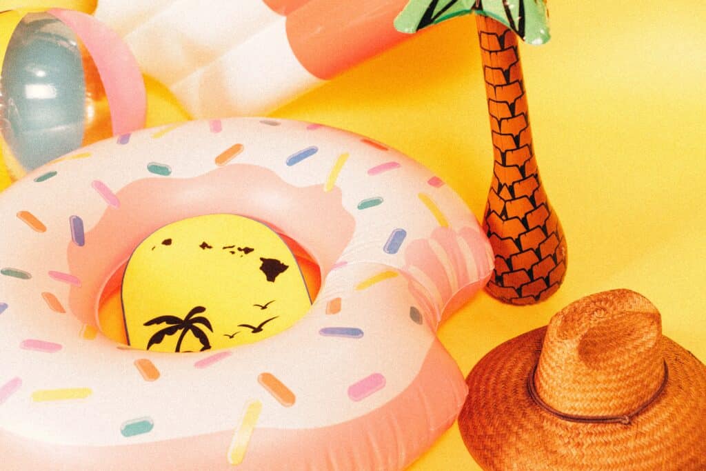 A pink doughnut-shaped pool float, a straw hat, an inflatable palm tree, and a beach ball are arranged on a yellow background, showcasing Summer Gifts To Suit Everyone with vibrant tropical and summery vibes.