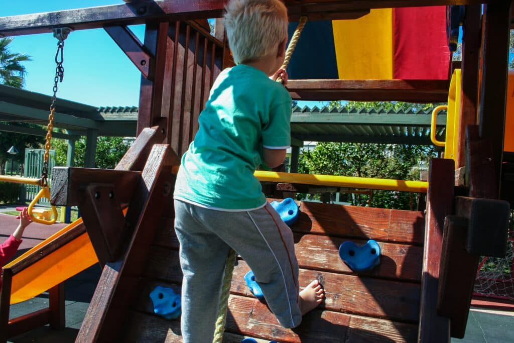 A young child with blond hair climbs a wooden play structure using blue climbing holds, outdoors on a sunny day. A yellow slide and rope are visible nearby in the playground, creating a fun space for children to play.