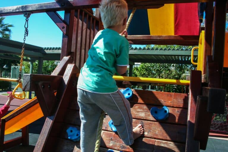 A young child with blond hair climbs a wooden play structure using blue climbing holds, outdoors on a sunny day. A yellow slide and rope are visible nearby in the playground, creating a fun space for children to play.