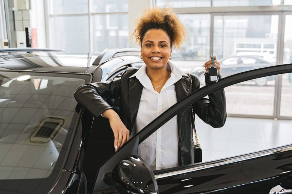 A smiling woman stands next to an open car door inside a showroom, holding a set of car keys—a reminder that smart decisions like this can be among the best investments parents can make for their family's future.