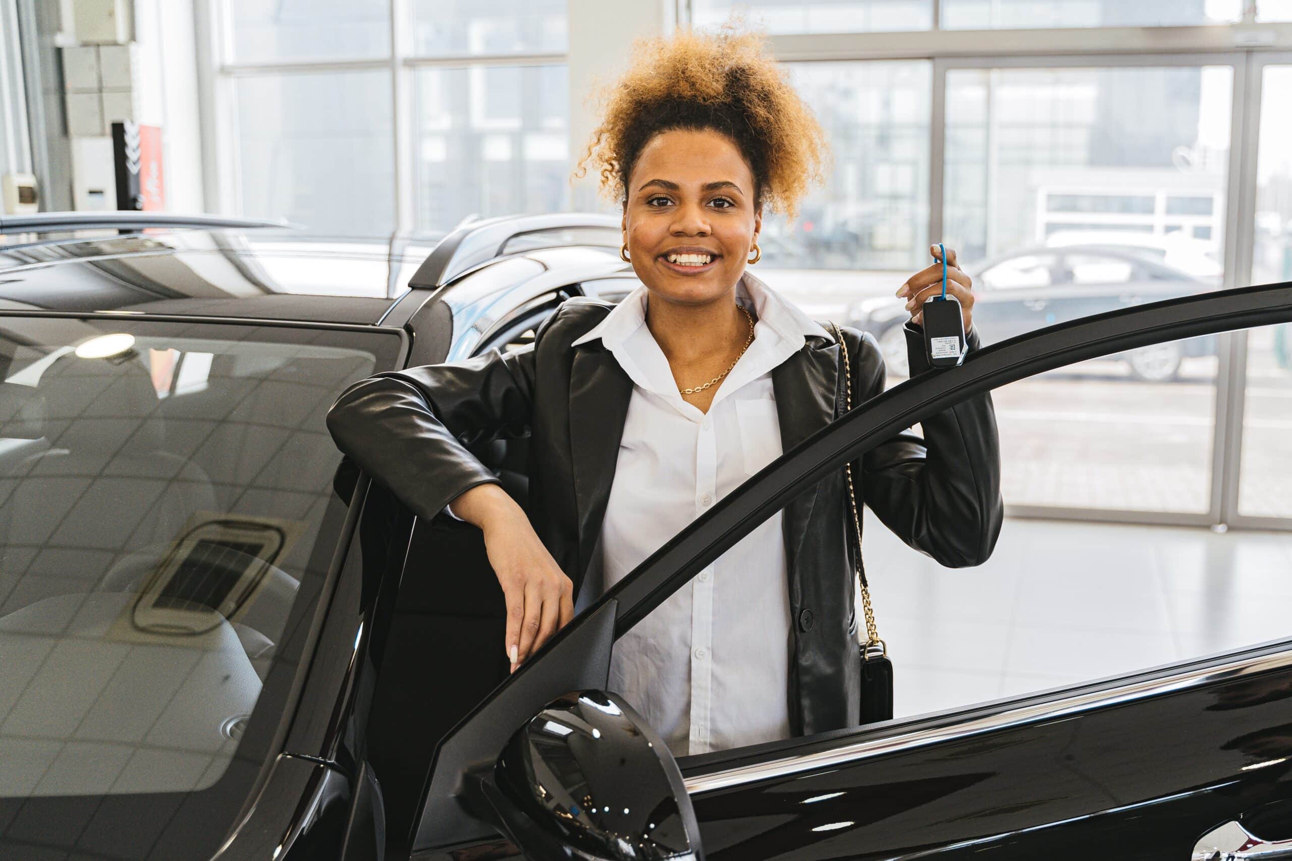 A smiling woman stands next to an open car door inside a showroom, holding a set of car keys—a reminder that smart decisions like this can be among the best investments parents can make for their family's future.
