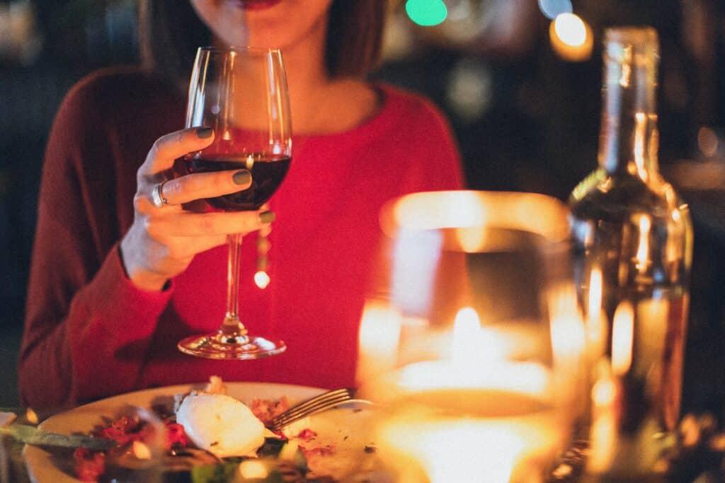 A person in a red top holds a glass of red wine at a candlelit dinner table, showing how to save money whilst still enjoying all the things you love, with blurred plates of food and a bottle in the background creating a warm, cosy atmosphere.