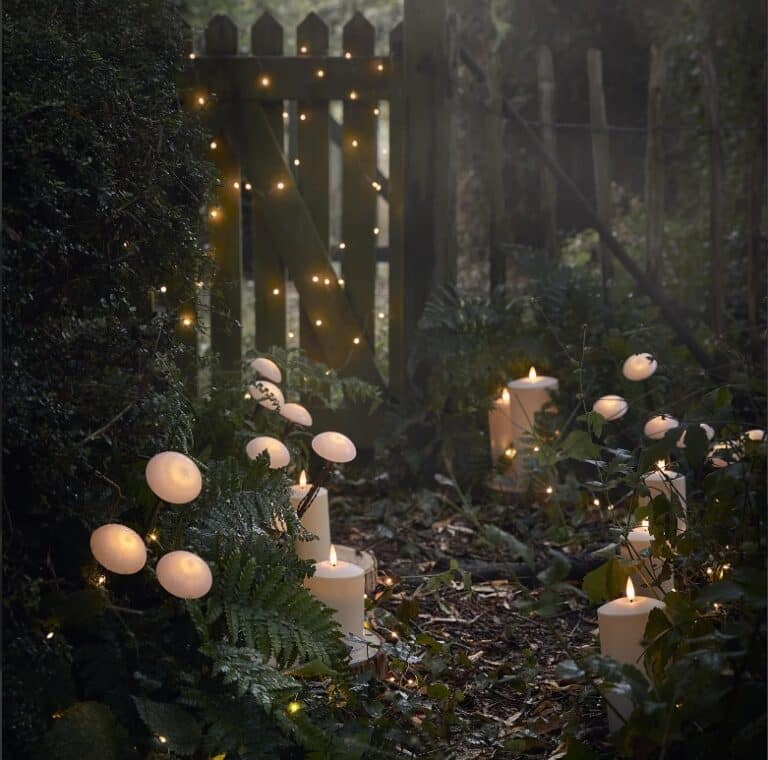 A woodland garden path is lit by white candles and glowing fairy lights, with eco-friendly, mushroom-shaped lights nestled among ferns and greenery, showing how to decorate your garden in an environmentally friendly way.