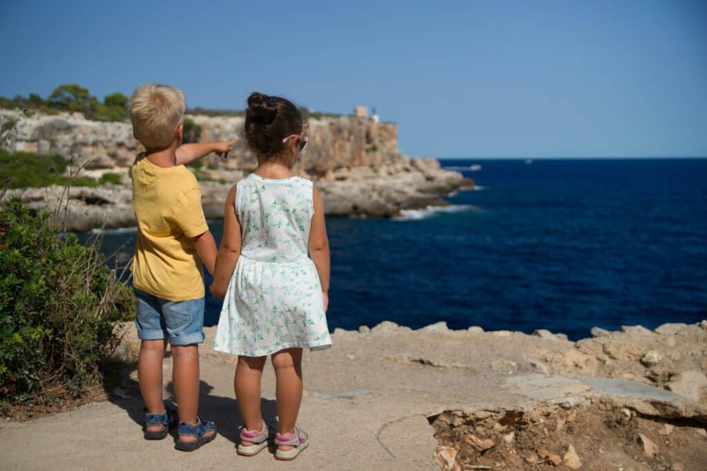 Two young children, a boy in a yellow shirt and a girl in a white dress, stand on a rocky path by the sea. The boy points towards the water and cliffs, sharing curiosity—an early lesson in how to raise a well-mannered child.
