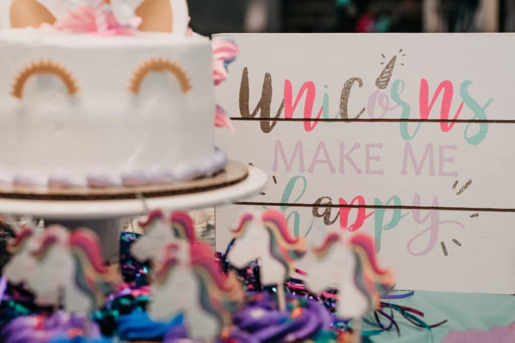 A unicorn-themed cake sits next to a colourful sign that reads “Unicorns make me happy,” surrounded by decorated unicorn treats, festive decorations, and playful Unicorn and Dinosaur toys in the foreground.