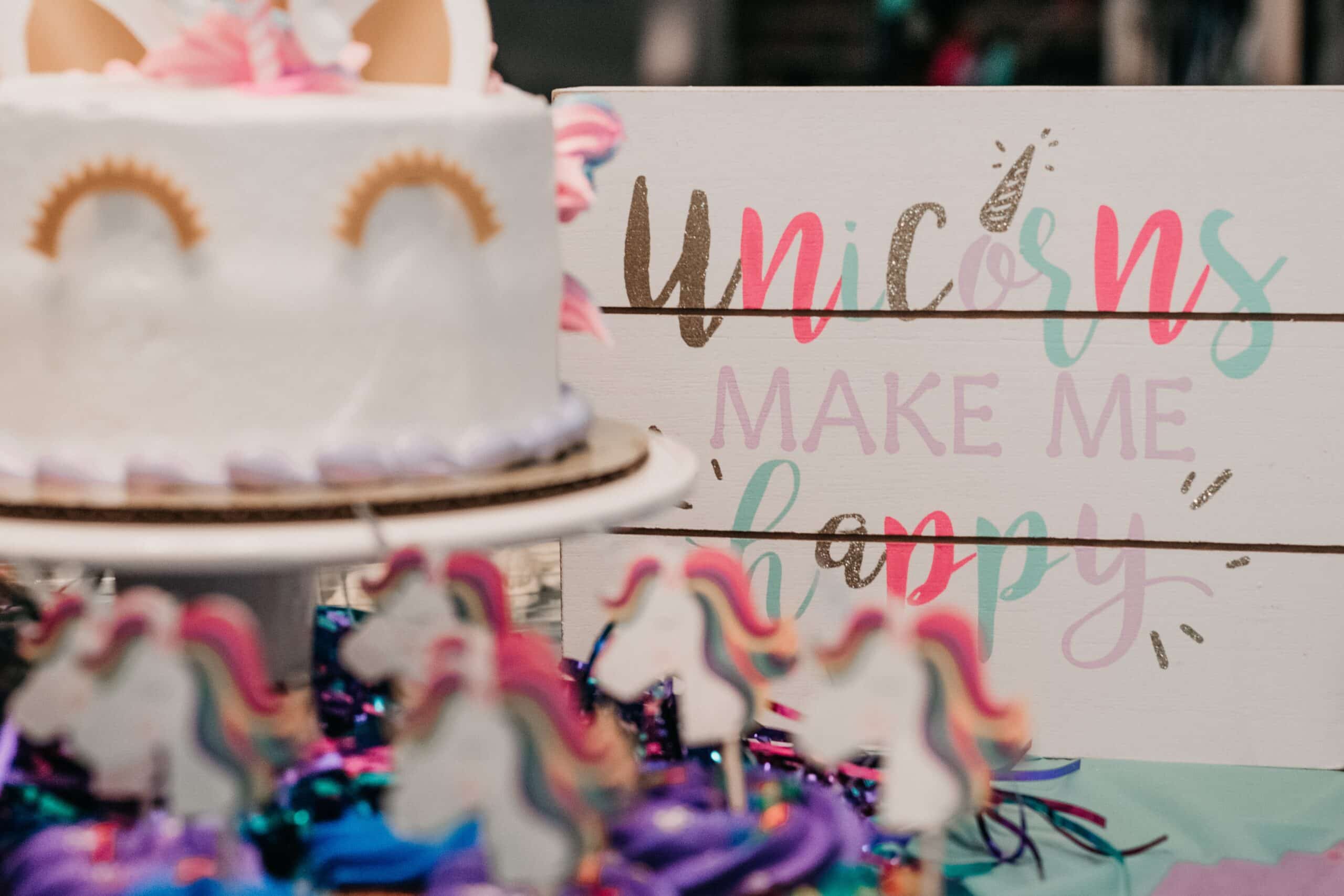 A unicorn-themed cake sits next to a colourful sign that reads “Unicorns make me happy,” surrounded by decorated unicorn treats, festive decorations, and playful Unicorn and Dinosaur toys in the foreground.