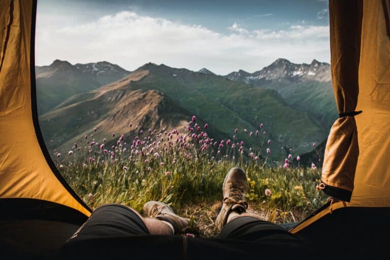 View from inside a tent, showing a person’s legs and feet facing a field of purple wildflowers with green mountains and a partly cloudy sky—perfect inspiration as you discover ways to get ready for your first solo trip abroad.