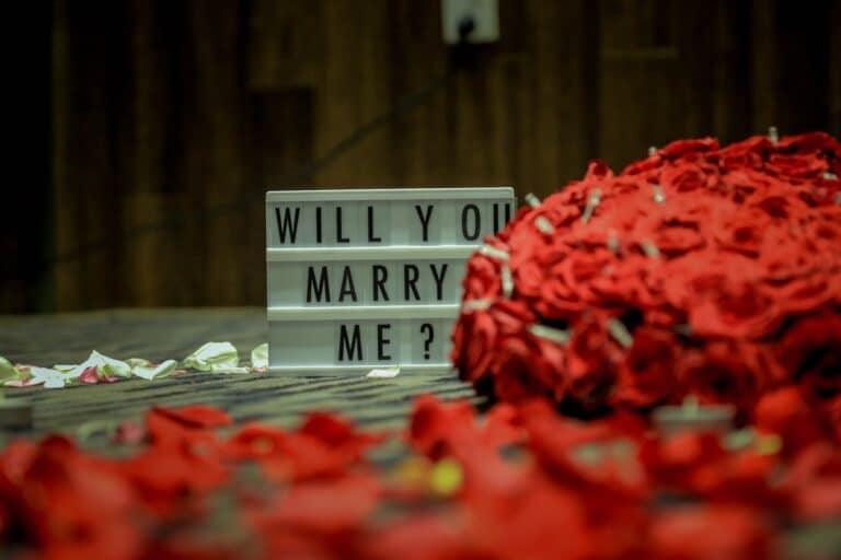 A lightbox sign reading "WILL YOU MARRY ME?" sits on the floor next to a large bouquet of red roses, scattered rose petals, and a box featuring unique engagement rings. The background is out of focus.