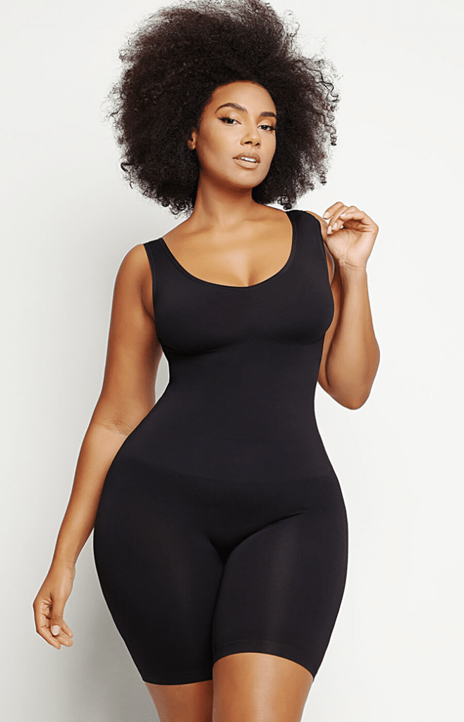 A woman with curly hair poses confidently wearing a fitted, sleeveless black bodysuit with mid-thigh length shorts, showcasing the latest trends and innovations in shapewear against a plain white background.