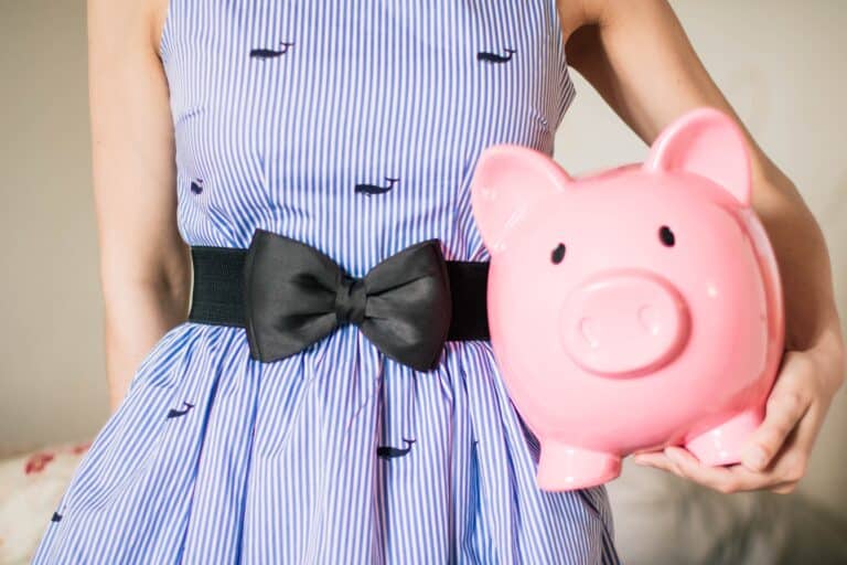 A person wearing a blue and white striped dress with a black bow belt is holding a pink piggy bank, symbolising How To Teach Children To Be Financially Responsible. The image is cropped to show only the torso and arm.