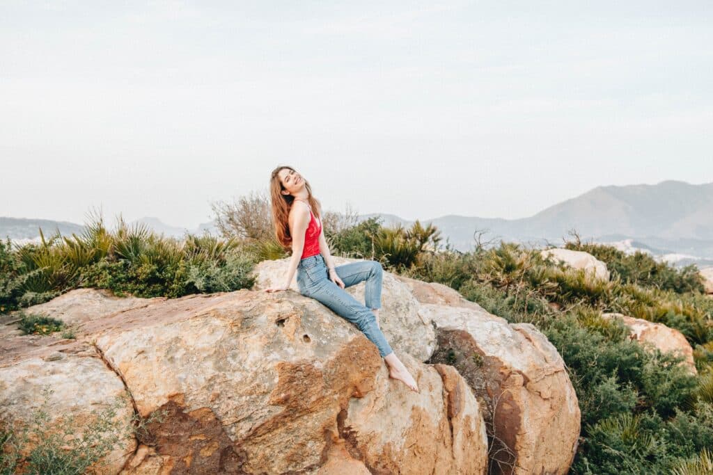 A woman in a red top and jeans sits barefoot on a large rock, surrounded by greenery and mountains under a pale blue sky, embracing the fantastic opportunities a gap year has to offer.