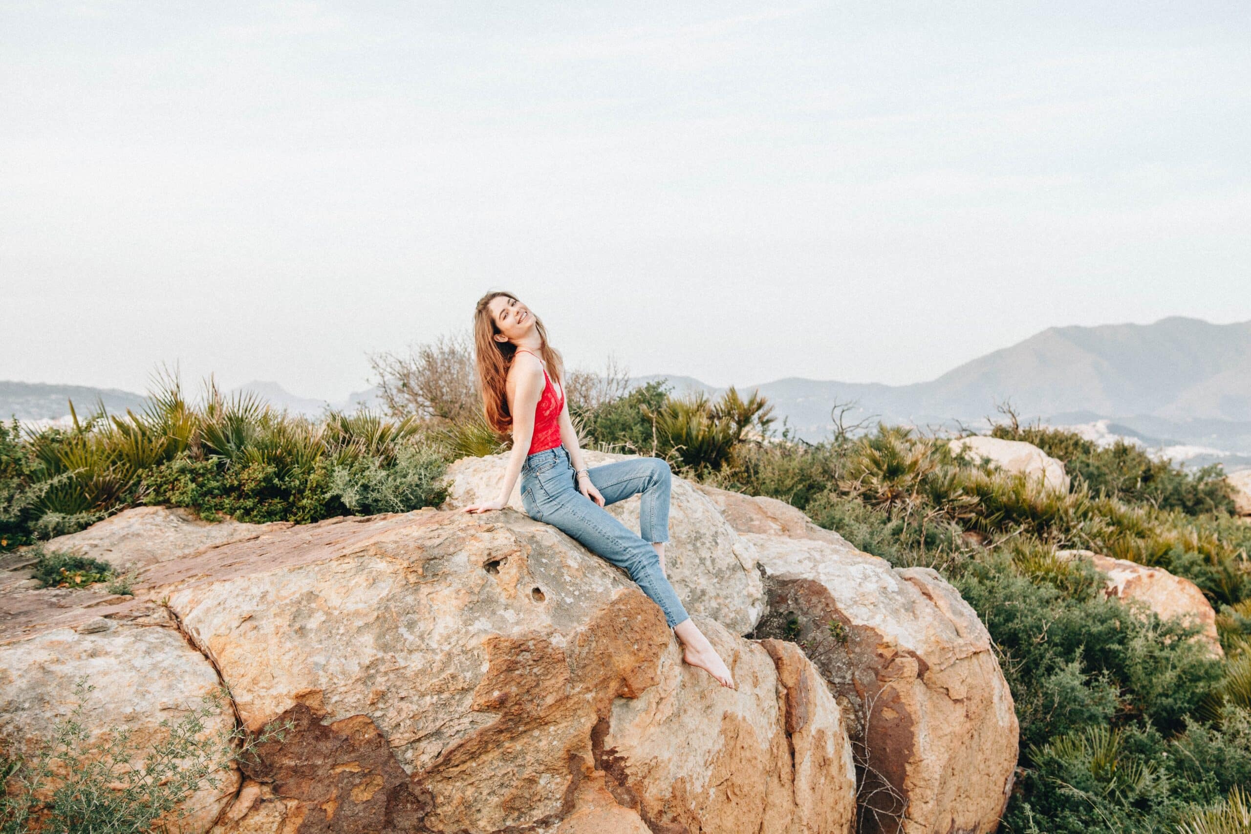 A woman in a red top and jeans sits barefoot on a large rock, surrounded by greenery and mountains under a pale blue sky, embracing the fantastic opportunities a gap year has to offer.