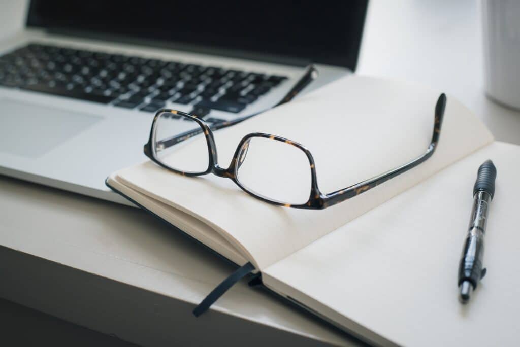 A pair of spectacles rests on an open notebook next to a black pen, with part of a laptop and a white mug visible in the background—an ideal setup for tackling Sole Trader Finances: Using an Accountant vs. Do-It-Yourself.